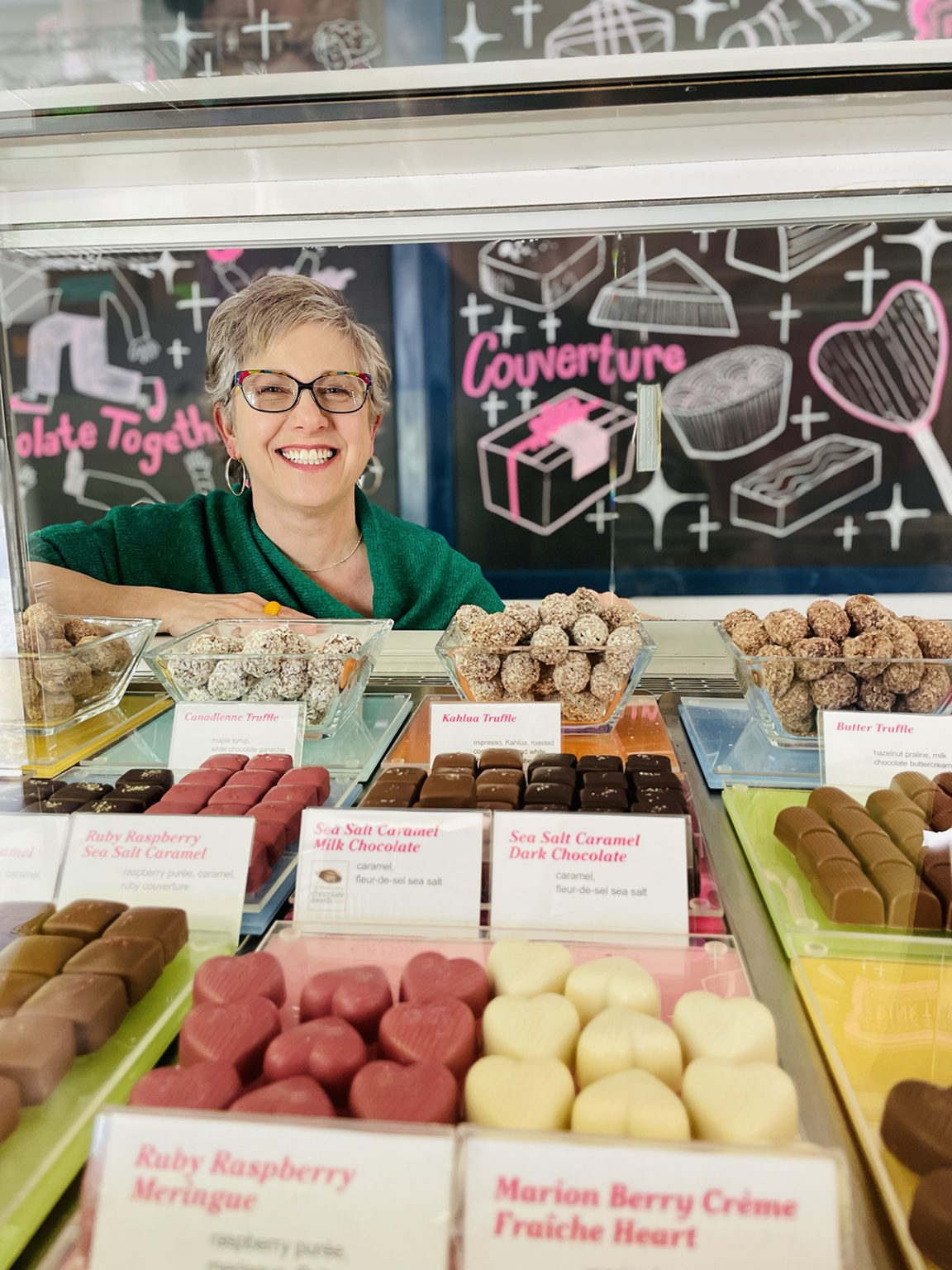 A smiling woman with grey hair and a green shirt stands in front of a stand with chocolates and confections.